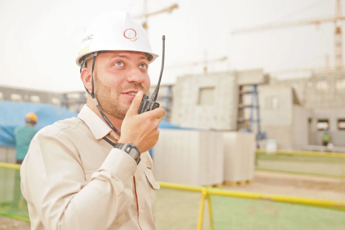 Construction site supervisor in a hard hat speaking into a handheld two-way radio in front of tower cranes