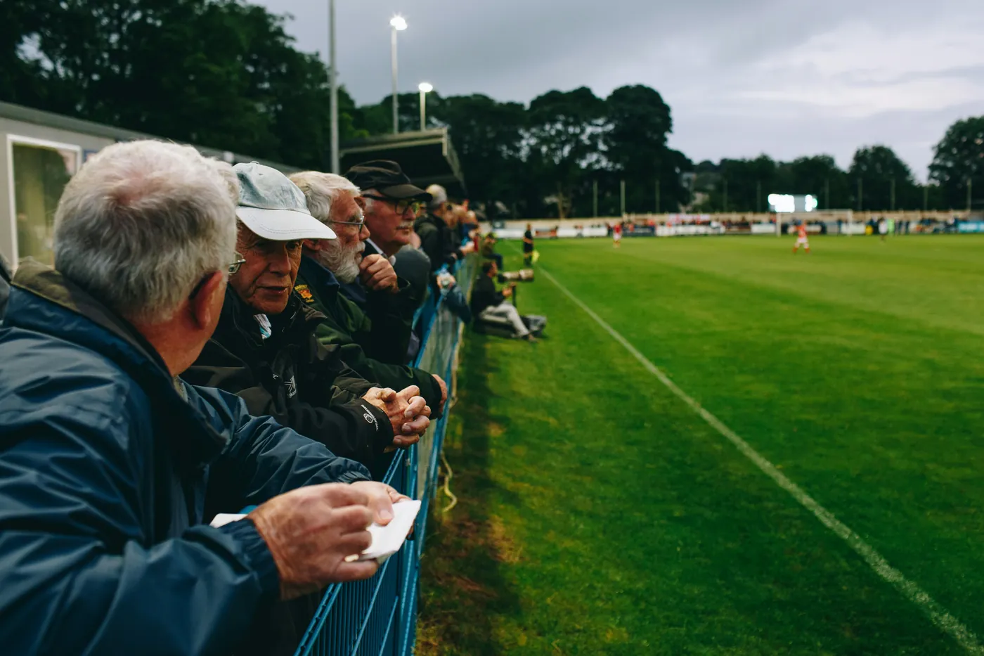 Spectators leaning on a pitch-side barrier watching a live football match at a sports ground.