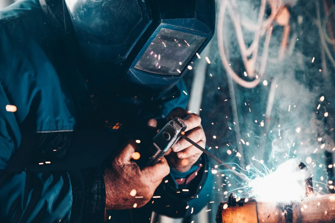 Welder in a protective visor striking an arc on a metal pipe with sparks flying, the kind of noisy industrial environment where UK teams depend on reliable push-to-talk radio communication