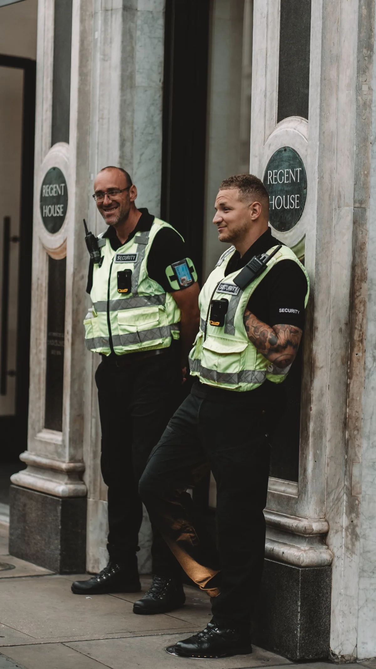 Two UK security officers in hi-vis vests with handheld two-way radios and body-worn cameras on duty outside the Regent House entrance
