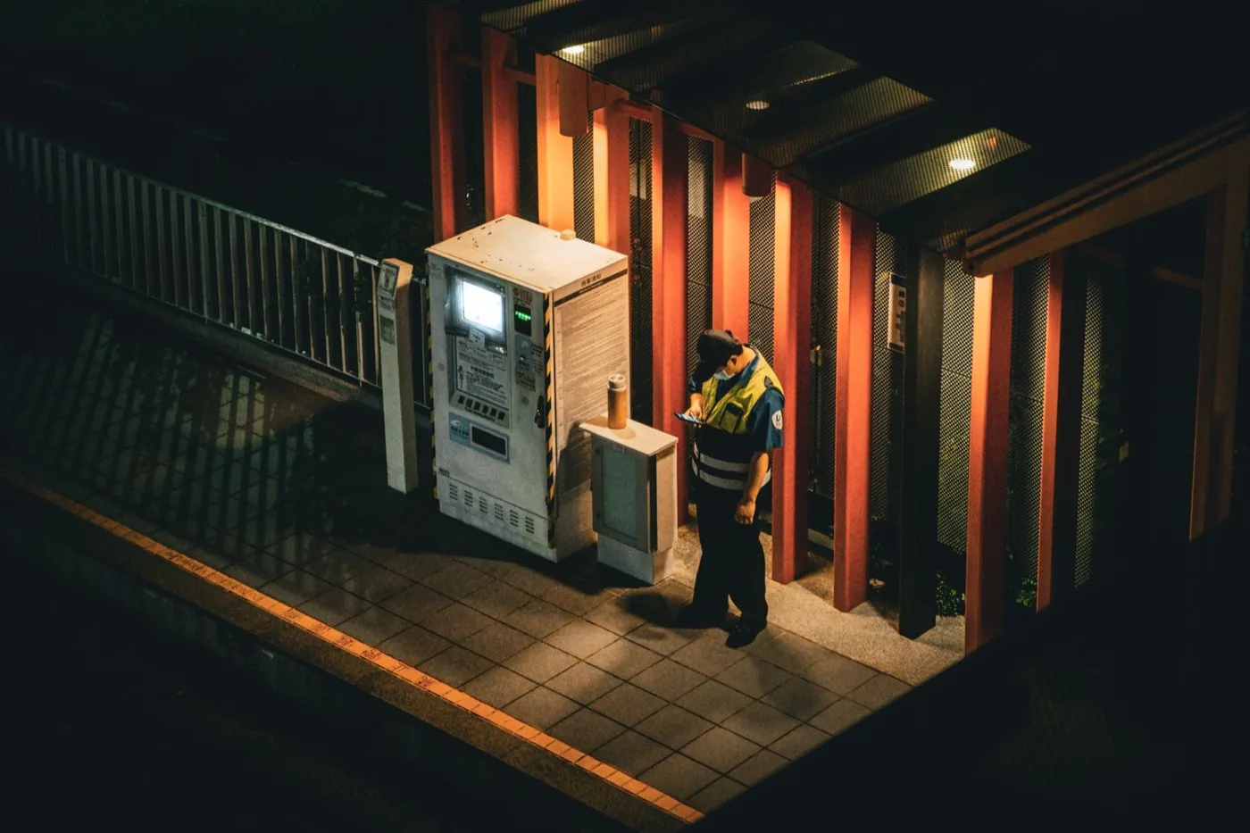 Security guard in a hi-vis vest working alone on a night shift, lit only by a ticket machine on a deserted platform